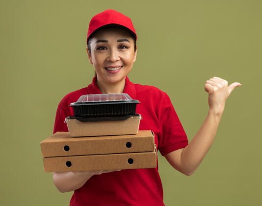 站着身穿红色制服 头戴鸭舌帽的年轻女送货员手拿比萨饼盒和食品包 友好地微笑着 手指着站在绿色墙壁上的一边帽子壁板女人