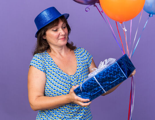 中间快乐快乐的中年妇女 戴着派对帽 手持一束五颜六色的气球 微笑着看着礼物 站在紫色的墙上庆祝生日派对年龄气球帽子
