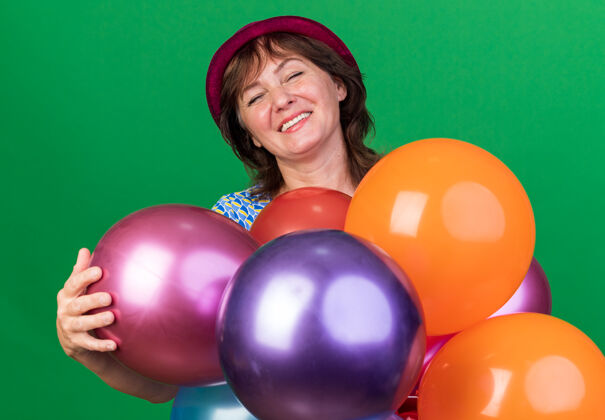 中间中年妇女戴着派对帽 手里拿着五颜六色的气球 站在绿色的墙上欢庆生日派对庆祝气球年龄