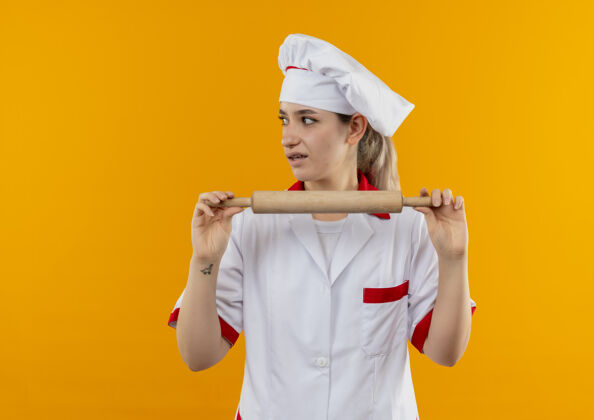 年轻年轻漂亮的厨师穿着厨师制服 手持擀面杖 看着橙色墙壁上孤立的一面 留着复印空间卷壁板烹饪