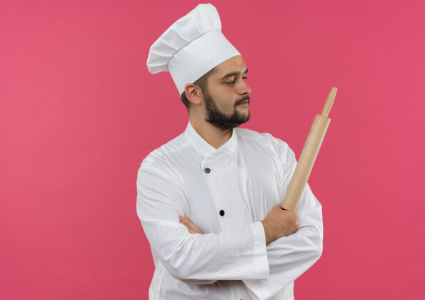 卷自信的年轻男性厨师身着厨师制服 站在那里 保持封闭的姿势 拿着擀面杖 看着粉色墙壁上孤立的一面 还有复制空间男性姿态信心