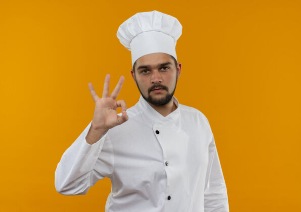 烹饪自信的年轻男性厨师身着厨师制服 在橙色的墙上做着“ok”的招牌 看起来很孤立 还有复印空间好的厨师制服