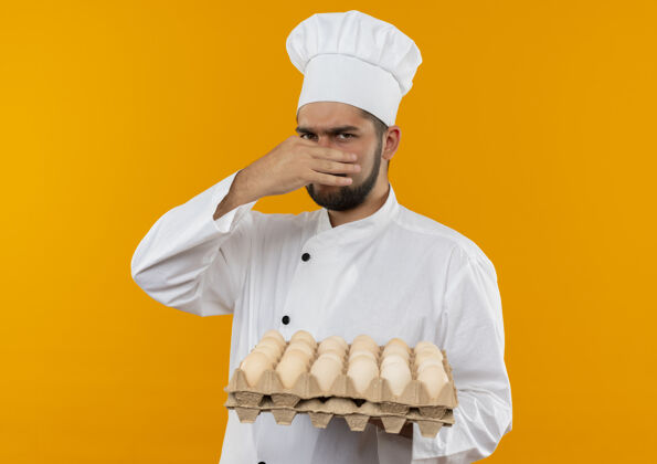 烹饪年轻的男厨师穿着厨师制服 手里拿着一盒鸡蛋 鼻子被隔离在橙色的墙上鼻子厨师纸箱