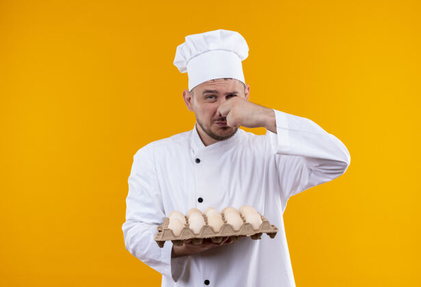帅气年轻帅气的厨师穿着厨师制服 手里拿着一盒鸡蛋 鼻子被隔离在橙色的墙上纸箱恼火烹饪