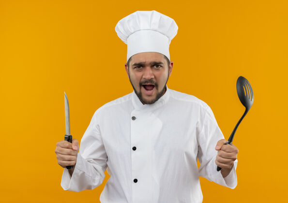 印象令人印象深刻的年轻男厨师在厨师制服举行刀和开槽勺子隔离在橙色墙上持有年轻勺子
