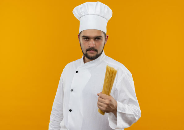持有自信的年轻男性厨师身着厨师制服 拿着意大利面 隔离在橙色的墙上 留有复制空间厨师意大利面年轻