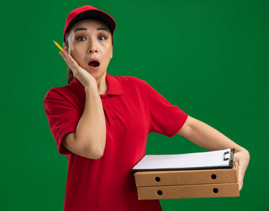 惊喜身穿红色制服 头戴鸭舌帽 拿着比萨饼盒和写字板 拿着钢笔的年轻女送货员站在绿色的墙上 既惊讶又惊讶帽子送货站着