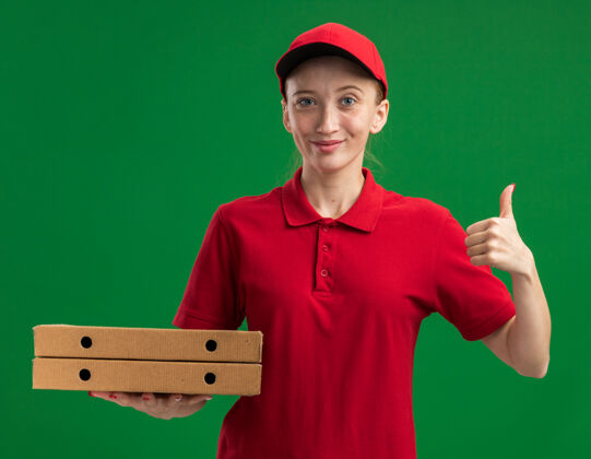 女孩身穿红色制服 头戴鸭舌帽 手拿披萨盒 自信微笑 竖起大拇指的年轻送货员站在绿色的墙上盒子送货自信