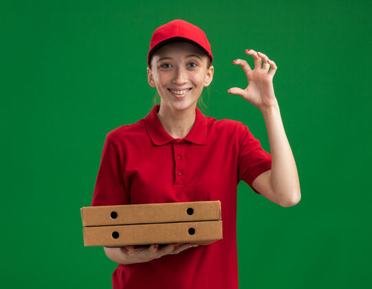 拿着身穿红色制服 头戴鸭舌帽 手拿披萨盒的年轻送货女孩站在绿色的墙上 做着一个小小的手势 手指欢快地微笑着微笑手势帽子
