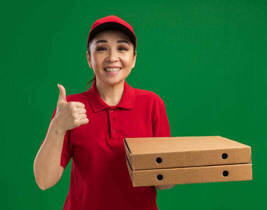 微笑身穿红色制服 头戴鸭舌帽 手持比萨饼盒 友好微笑 竖起大拇指的年轻送货员站在绿色的墙上站着制服朋友