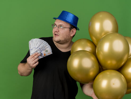 印象令人印象深刻的成年斯拉夫人戴着眼镜 戴着蓝色的派对帽 手里拿着氦气球和钱在一旁看着光学穿绿色