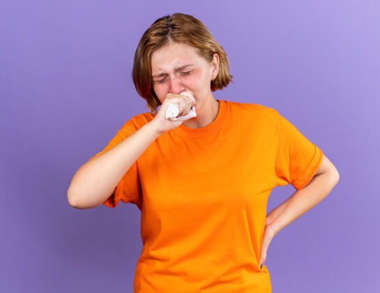 立场不健康的年轻女子穿着橘色t恤感觉可怕的咳嗽拳头患有病毒站在紫色的墙壁拳头咳嗽年轻