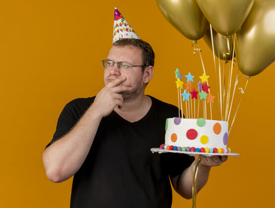 男人体贴的成年斯拉夫人戴着眼镜 戴着生日帽 手放在下巴上 手里拿着氦气球和生日蛋糕 看着身边手气球复制