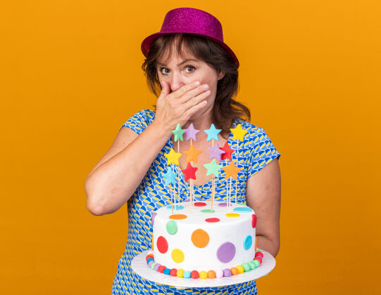 年龄中年妇女戴着派对帽拿着生日蛋糕被震得捂着嘴用手庆祝生日派对站在橙色的墙上蛋糕生日封面
