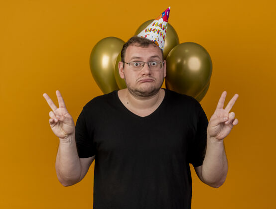 光学震惊的成年斯拉夫男子戴着眼镜 戴着生日帽 站在氦气球前 用双手示意胜利手势标志手势站