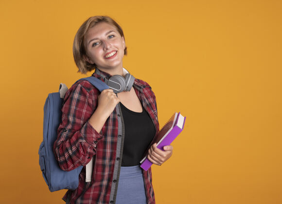 抱着微笑的年轻斯拉夫女学生戴着耳机 背着背包 站在一边拿着书和笔记本耳机年轻人背包