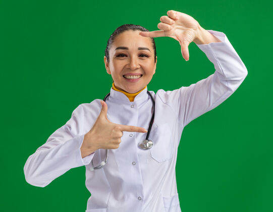 外套快乐积极的年轻女医生 穿着白药衣 脖子上戴着听诊器 手指站在绿色的墙上做着手势使女人积极