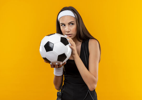 腕带自信的年轻漂亮的运动女孩戴着头带和腕带拿着足球看一边跳绳围着她的脖子孤立在橙色的墙上壁板周围头带