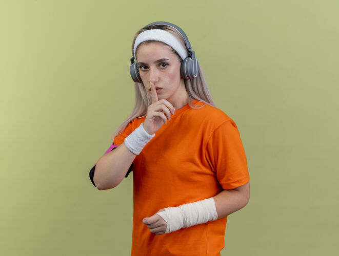 绿色自信的年轻白人运动女孩戴着牙套戴着耳机戴着头带腕带和手机臂带手势沉默的标志臂带耳机运动