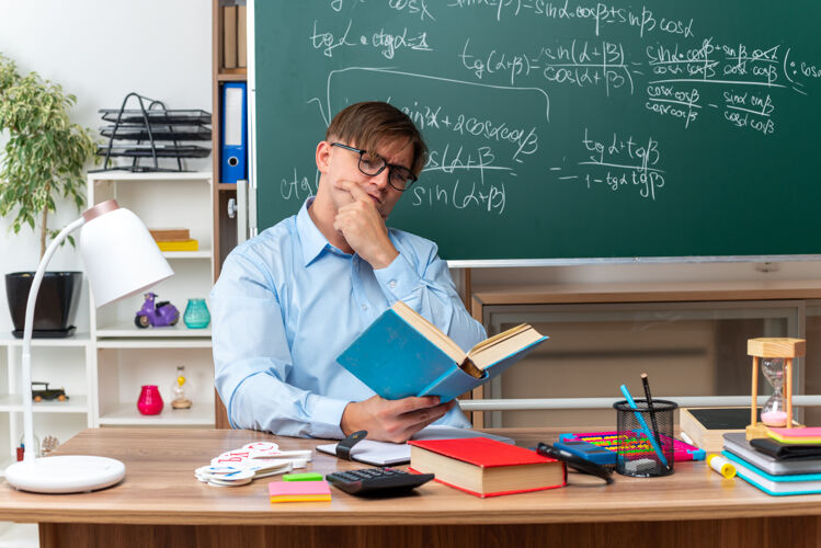黑板年轻的男老师戴着眼镜认真看书备课坐在课桌旁 教室里黑板前放着书和笔记坐着课人