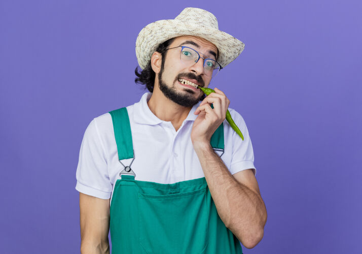 穿年轻的留着胡须的园丁 穿着连身衣 戴着帽子 站在蓝色的背景上 脸上露出绿色的辣椒色 脸上露出困惑的微笑男人胡须花园