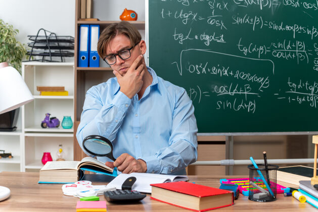 放大镜年轻的男老师戴着眼镜坐在书桌旁 拿着书和笔记 透过放大镜看着教室黑板前严肃的书本学校玻璃穿过