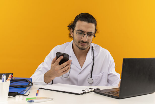 办公桌年轻的男医生戴着医用眼镜 穿着医用长袍 听诊器坐在办公桌旁电话长袍工具