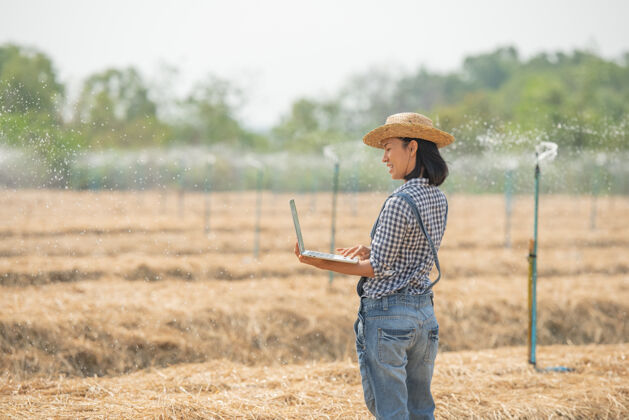 互联网亚洲年轻的女农民戴着帽子站在地里 在笔记本电脑键盘上打字带着笔记本电脑的妇女监督农田 概念生态 交通 清洁空气 食品 生物制品的工作人生态工人