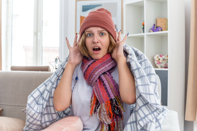 坐着不健康的年轻女子 戴着暖和的帽子 裹着毯子 看起来不舒服 生病 患感冒和流感 体温计发烧 坐在明亮的客厅里的沙发上 看起来又担心又害怕感冒房间不健康