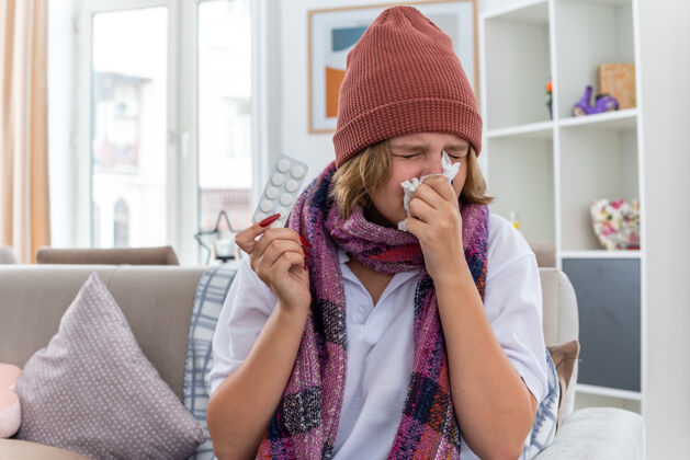 房间不健康的年轻女子戴着暖和的帽子 戴着围巾 用纸巾擤鼻涕 打喷嚏 患感冒和流感 坐在明亮的客厅椅子上年轻围巾纸巾