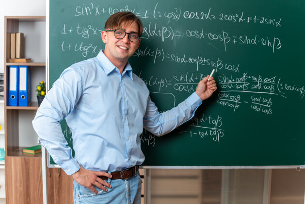 黑板年轻的男老师戴着眼镜拿着粉笔在教室里用数学公式站在黑板旁边讲解功课老师年轻人拿着
