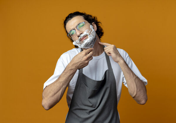 剃刀一个年轻的白人理发师 戴着眼镜 穿着制服 用剃须刀刮胡子 脸上涂着剃须膏 手指放在脖子上空间年轻奶油