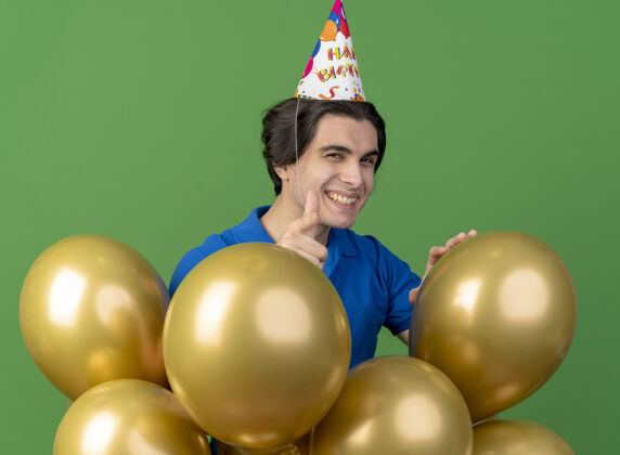 气球高加索帅哥戴着生日帽微笑着站着 氦气球指着摄像机空间氦绿色