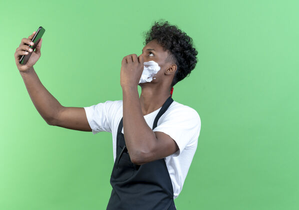 面部站在侧视图年轻的非洲裔美国男理发师穿着制服 脸上涂着剃须膏自拍绿色穿站