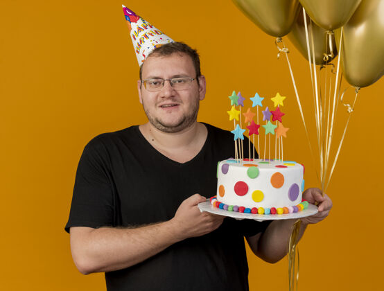 男人高兴的成年斯拉夫男子戴着眼镜戴着生日帽手持氦气球和生日蛋糕看着相机穿斯拉夫复制