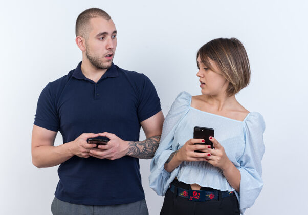 男朋友年轻漂亮的情侣男女拿着智能手机困惑的女人看着可疑的男友智能手机站着漂亮情侣站着