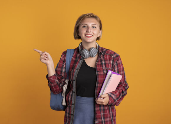 拿着微笑着的年轻斯拉夫女学生戴着耳机 背着书包 拿着笔记本指着身边复制女孩空间