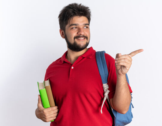 胡子快乐和积极的年轻留着胡子的学生 穿着红色马球衫 背着背包 拿着笔记本 看着一边 用食指指着一边站着笔记本背包站着