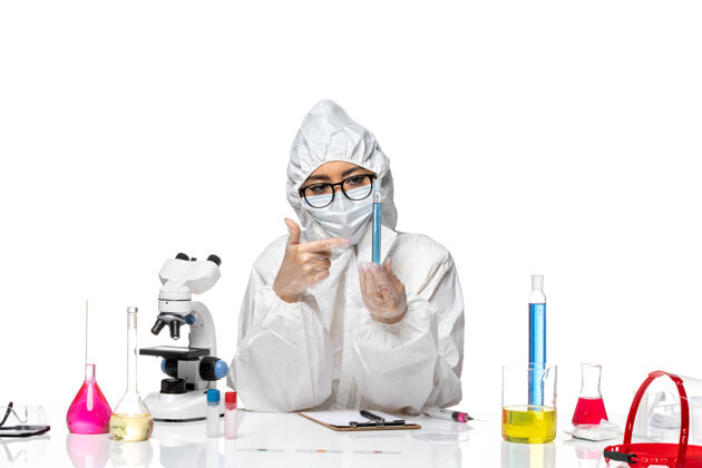 防护正面图身穿特殊防护服的年轻女化学家拿着放在白色桌子上的蓝色溶液烧瓶-化学病毒实验室病毒溶液套装