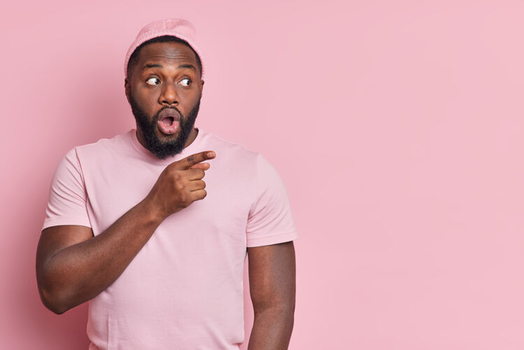 成人摄影棚拍摄的震惊的留着胡须的非裔美国男子站在旁边的复制空间显示 一些令人难以置信的穿着随意的广告对象隔离在粉红色的墙壁皮肤指点情感