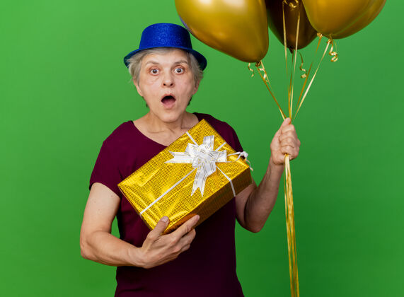 聚会震惊的老妇人戴着聚会帽 手里拿着氦气球和礼品盒上的绿色女人氦老人