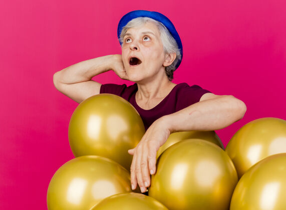 老人惊讶的戴着派对帽的老妇人把手放在头上 身后站着氦气球帽子粉色头