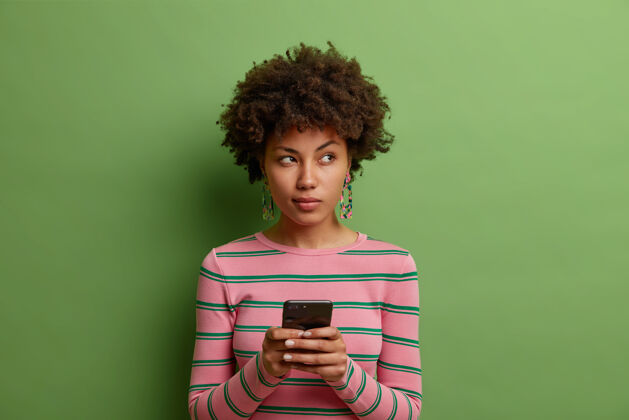 成人沉思的非裔美国妇女在智能手机上使用约会应用程序 目光远去穿着休闲毛衣隔着绿墙考虑电话卷曲
