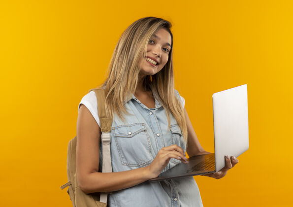 微笑微笑着的年轻漂亮的女学生 背着书包 拿着笔记本电脑 隔离在橙色的墙上女孩人学生