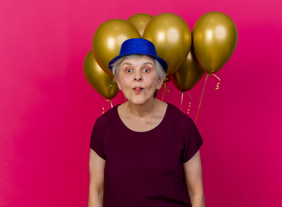 女人戴着派对帽的快乐的老妇人站在氦气球前结结巴巴地看着粉红色的相机前面舌头欢乐