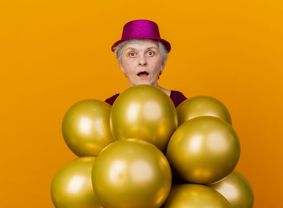 惊喜惊讶的老妇人戴着派对帽站在橙色的氦气球上氦帽子女人