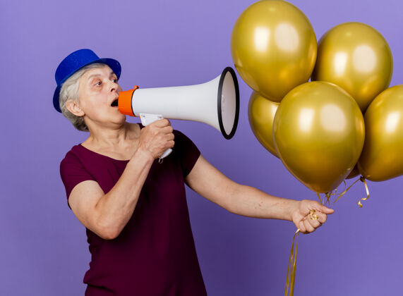 惊喜一位戴着派对礼帽的惊讶的老妇人手持氦气球 对着扩音器讲话壁板演讲者说话