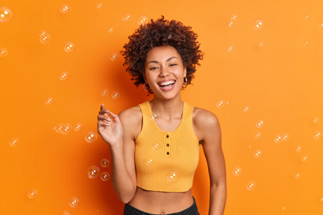 乐趣年轻的美国黑人少女 面带微笑 表情悠闲 穿着休闲服 在橙色的墙上摆出肥皂泡美丽肖像人