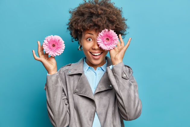 人积极的女人卷发用粉色非洲菊遮住眼睛黛西穿着灰色夹克和衬衫隔着蓝色的墙用两朵粉色的花遮住了美丽的眼睛女人头发夹克