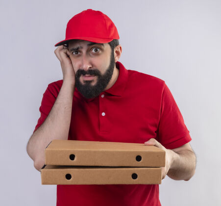 交货体贴的年轻帅气的送货员 穿着红色制服 戴着帽子 手里拿着披萨盒 笔直地看着 把手放在隔离在白墙上的脸颊上表情盒子人